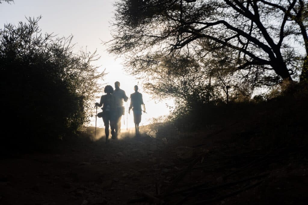 Three walkers silhouetted in dust and low sun as they set out from camp at dawn